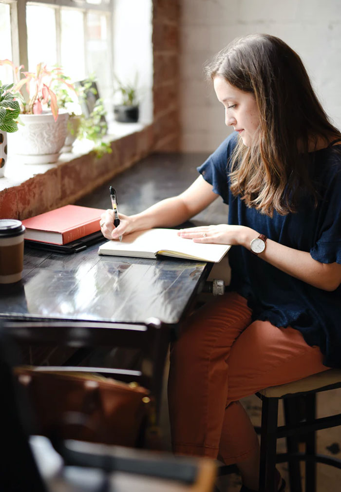 Woman sitting at a table by the window, writing in a notebook with books and coffee nearby, illustrating things easier if you're a woman.