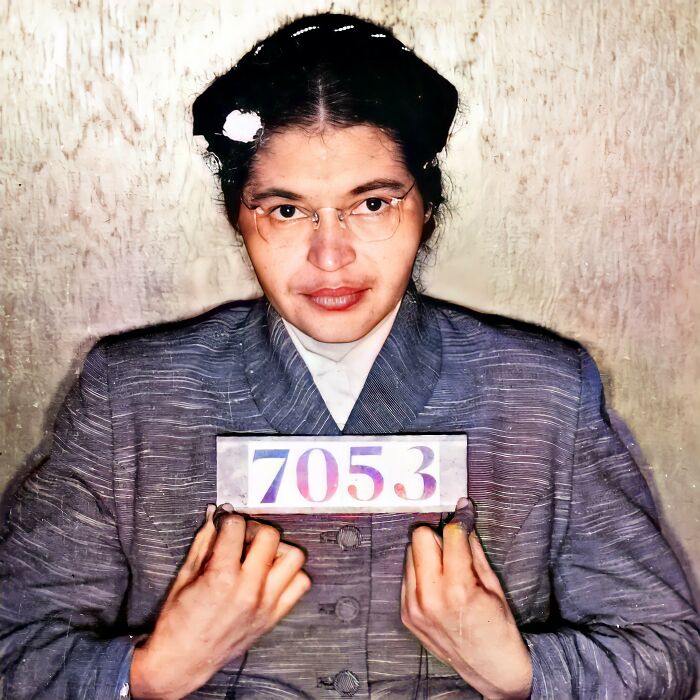 Restored vintage mugshot of a famous person holding a numbered plaque in a gray suit against a plain background.