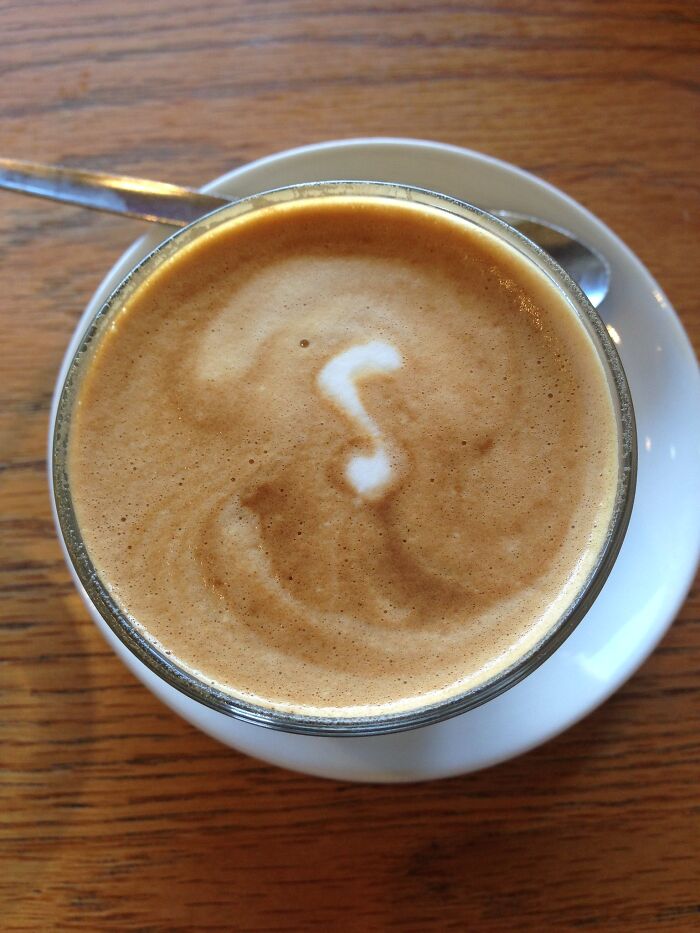 Close-up of coffee with foam art resembling a musical note on a wooden table with a spoon and saucer.
