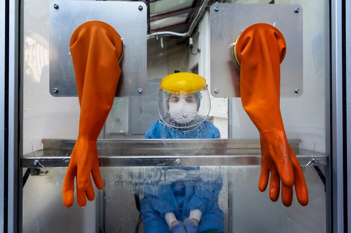 Medical professional in protective gear inside testing booth with orange gloves, featured in powerful Siena International Photo Awards.