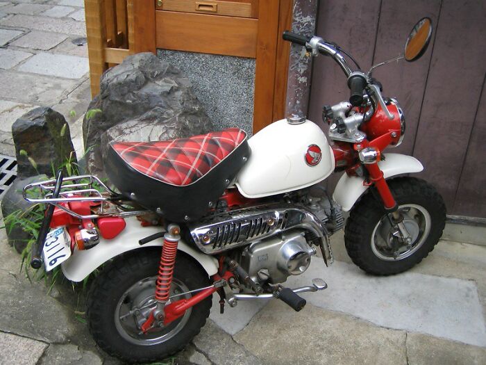 Small vintage motorcycle with red and white paint, parked beside a wooden wall and stone pavement outdoors.