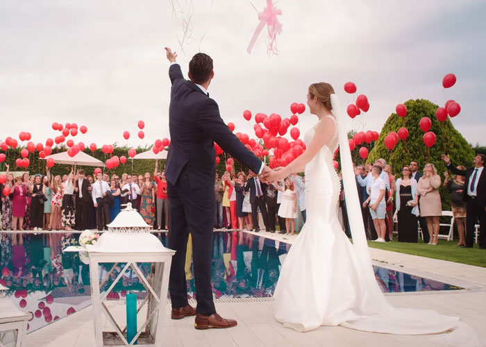 Newlywed couple releasing red balloons by pool surrounded by guests, illustrating scams in daily lives concept.