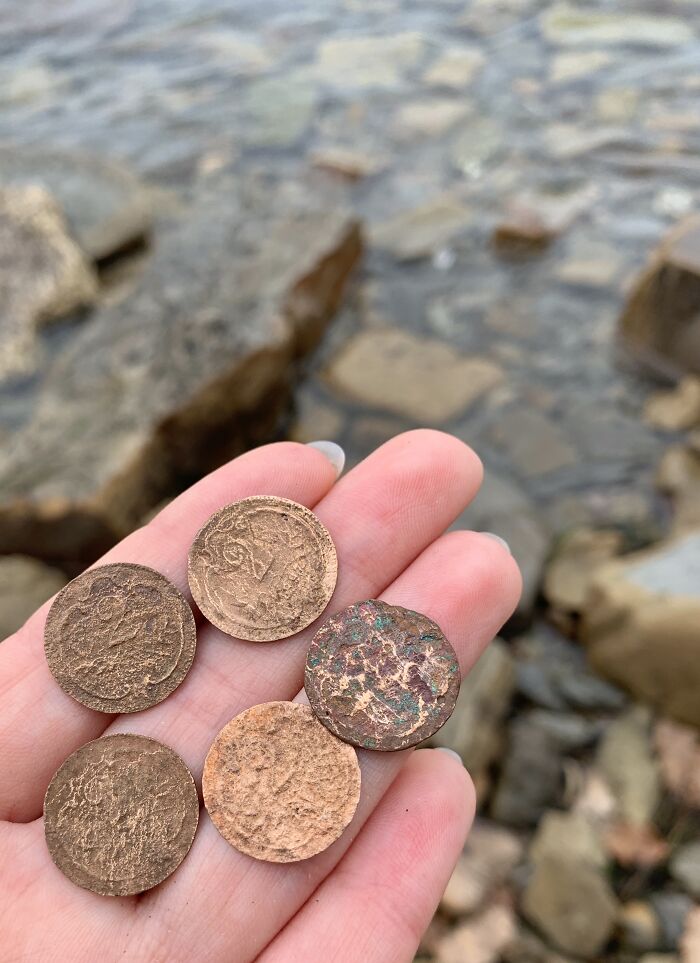 Hand holding five antique coins found as beach treasure with rocky shoreline and water in the background.