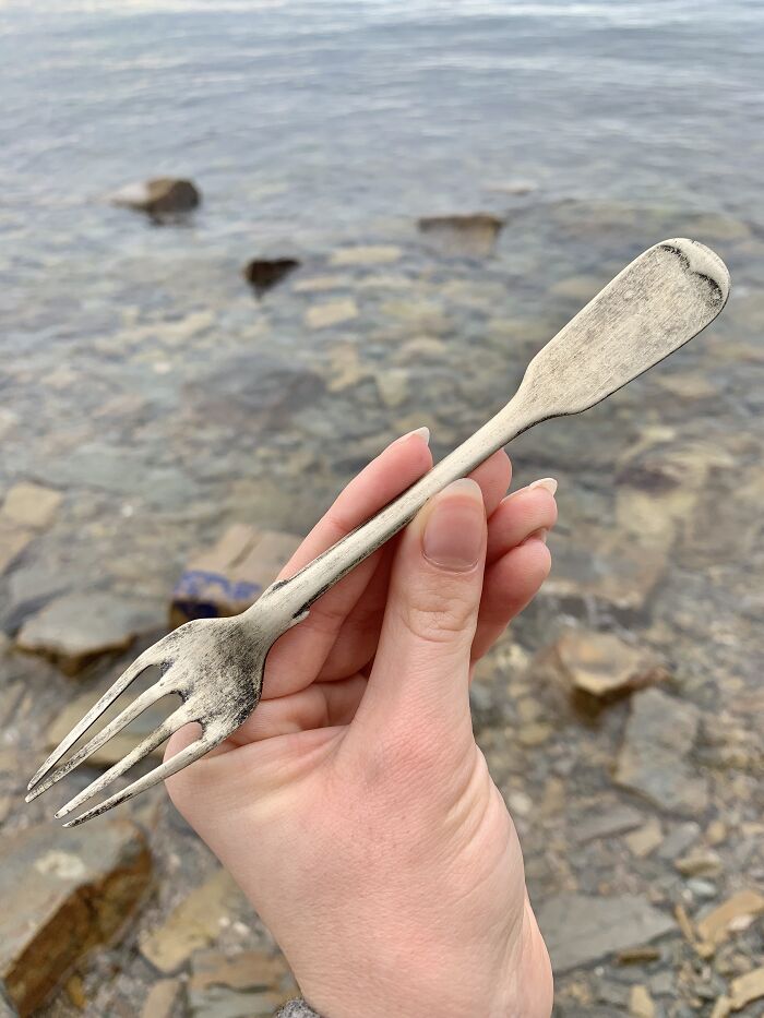 Hand holding an antique tarnished fork found as treasure washed up on the beach near rocky shoreline.