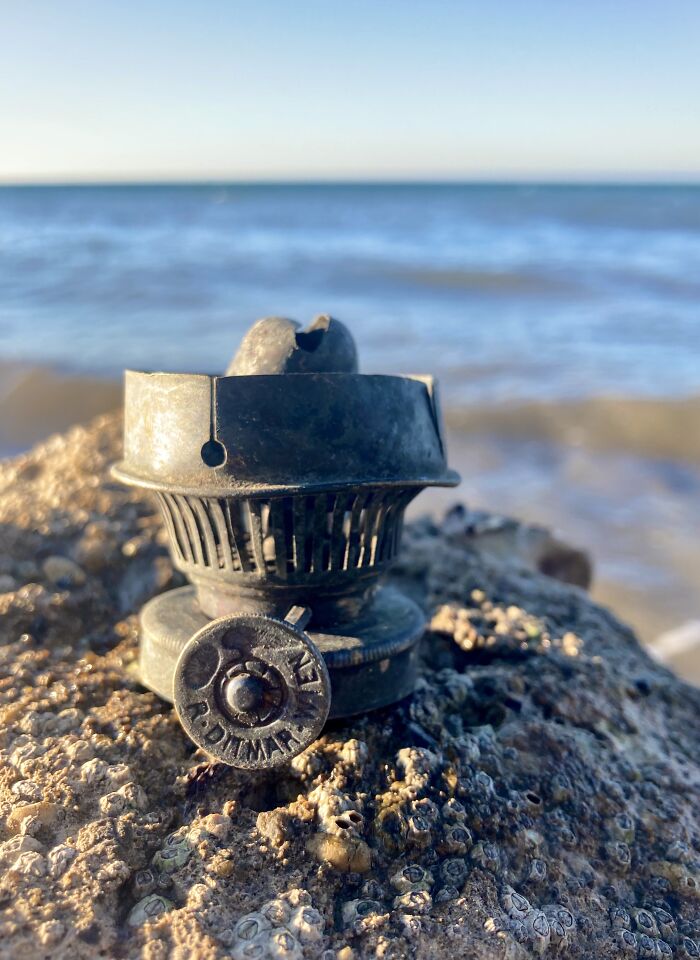 Antique metal object found on a rocky beach with ocean waves in the background, a unique treasure washed ashore.