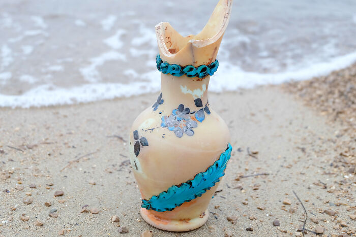 Antique vase with blue floral designs found washed up on the beach shore among sand and pebbles.