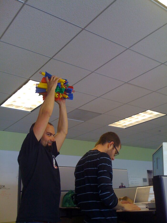 Two men in an office, one holding colorful plastic tubes overhead, sharing advice in an online group for young adults.