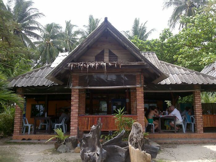 Rustic tropical outdoor dining area with people seated, surrounded by palm trees and greenery on a sunny day.