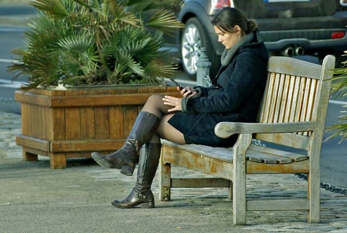 Woman sitting on a wooden bench outdoors, wearing boots and coat, focused on her smartphone in a public space.