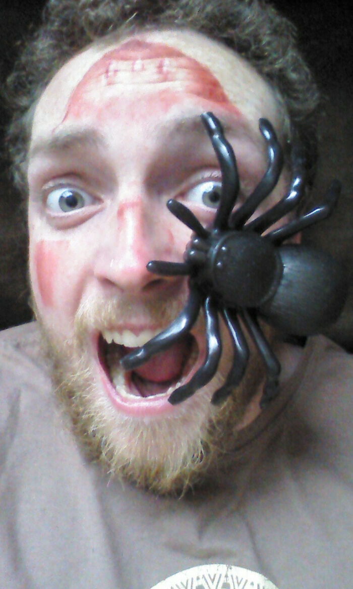 Man with simple Halloween costume featuring fake spider on face and minimal makeup, showing a surprised expression