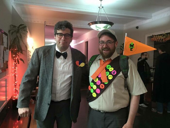 Two men in simple Halloween costumes, one dressed as a formal character and the other as a scout with badges and a flag.