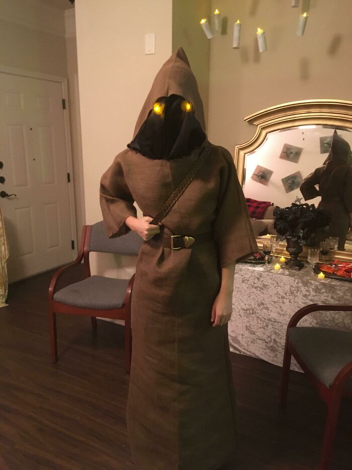 Person in a simple Halloween costume with glowing eyes wearing a brown hooded robe and black face covering indoors.