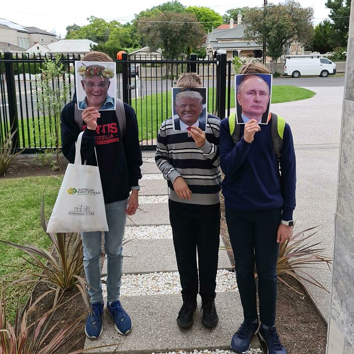Three people holding printed celebrity face masks as simple Halloween costumes outdoors on a sidewalk pathway.