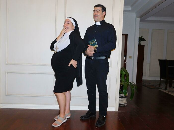 Couple with simple Halloween costumes dressed as a pregnant nun and a priest holding a Bible indoors.