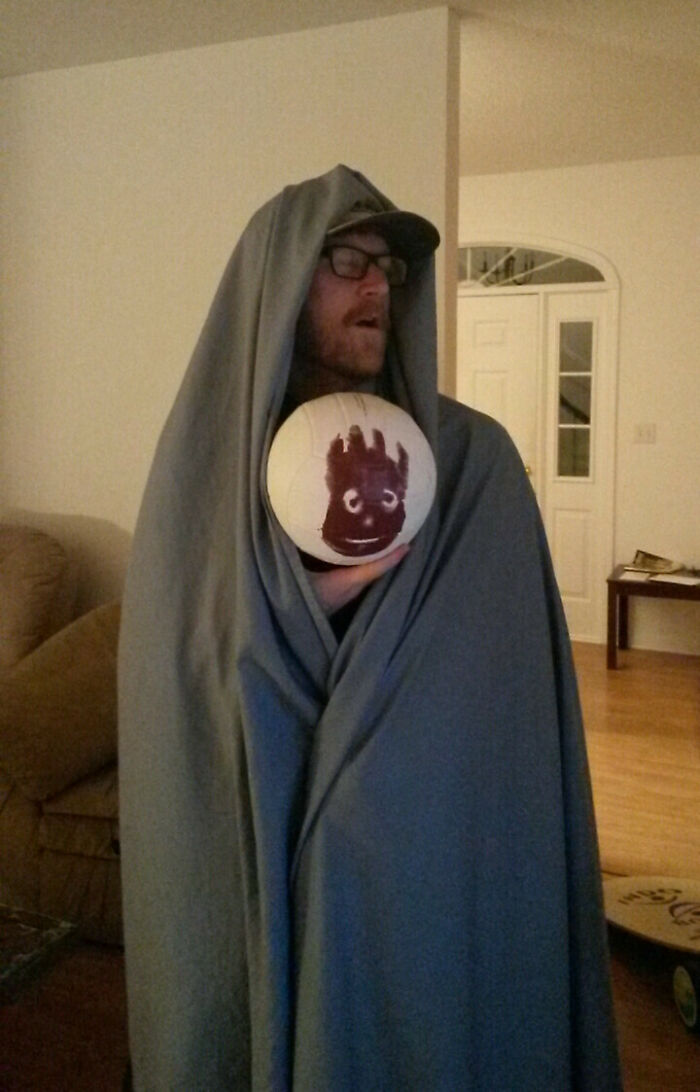 Man wrapped in gray blanket holding volleyball with a drawn face, a creative Halloween costume with minimal effort.