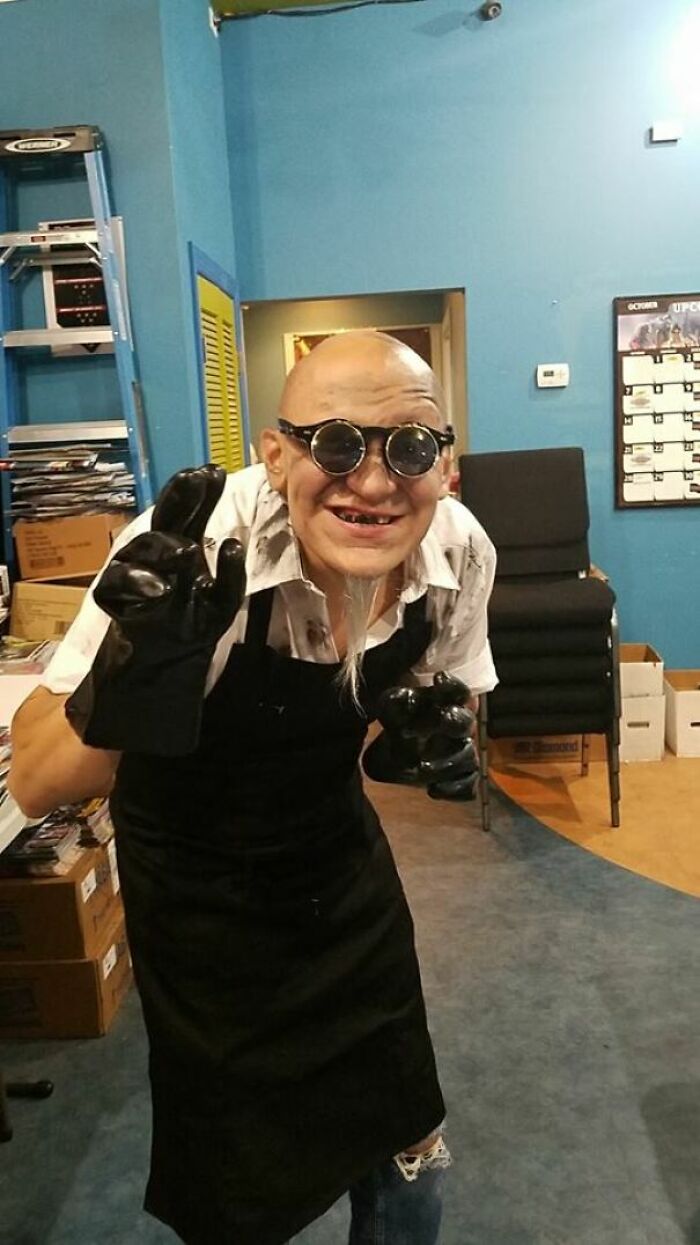 Person in a simple Halloween costume with round glasses, black gloves, and apron posing in an indoor setting.