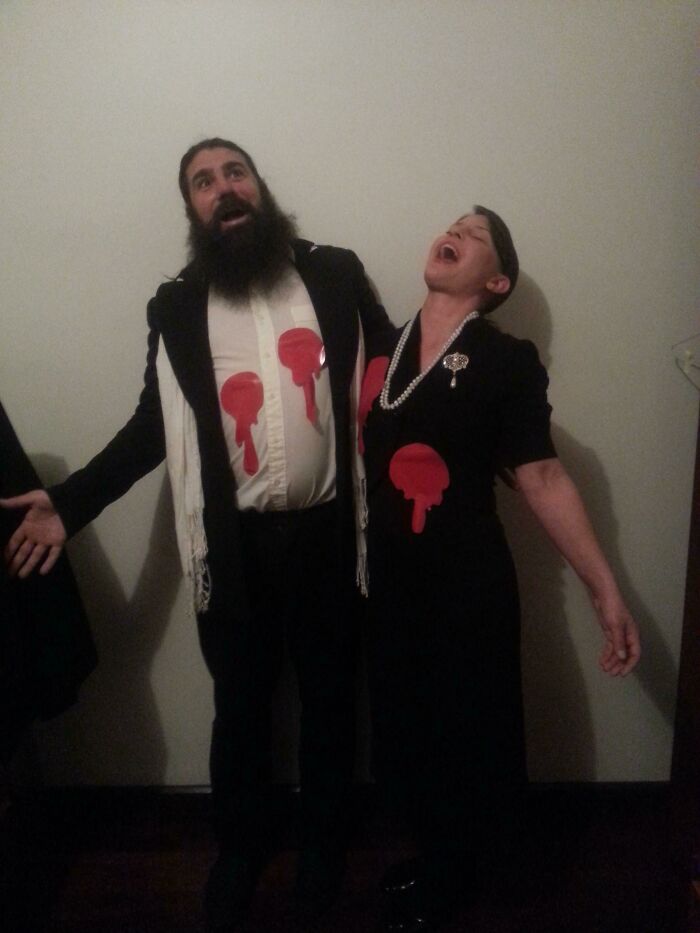 Couple wearing simple Halloween costumes with red paint splatters, laughing and posing indoors for a casual photo.