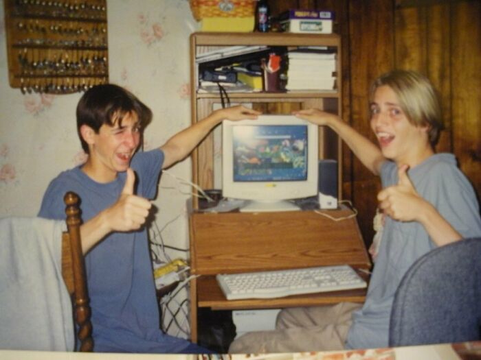 Teens with thumbs up by an early 2000s computer, showcasing nostalgic tech and fashion.