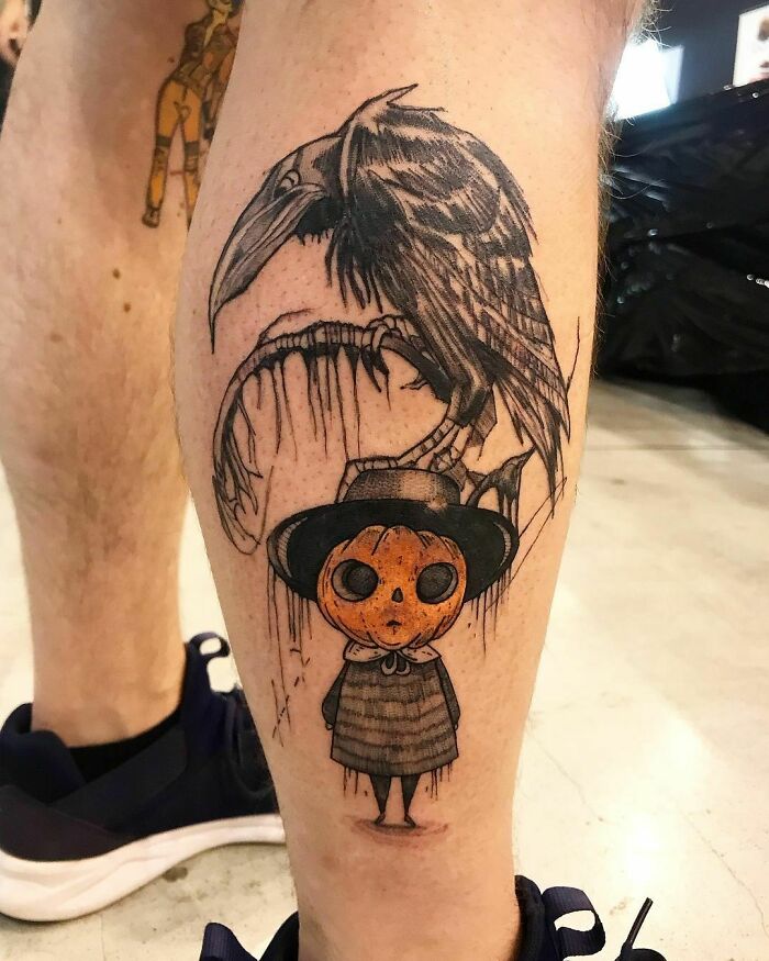 Spooky Halloween tattoo on leg featuring a raven and cartoonish character with an orange pumpkin head.