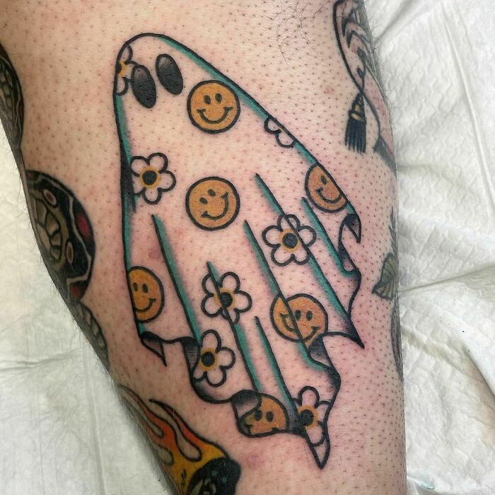 Spooky Halloween tattoo of a ghost with smiley faces and flowers, done on a person's leg.