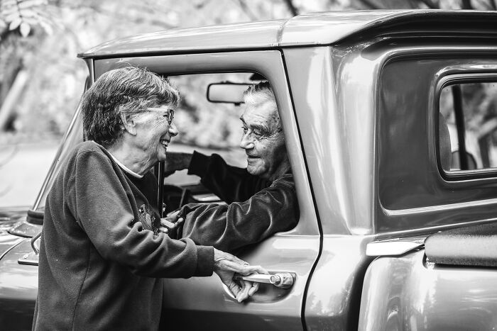 Elderly couple in love smiling warmly at each other by a vintage car in a candid romantic moment.