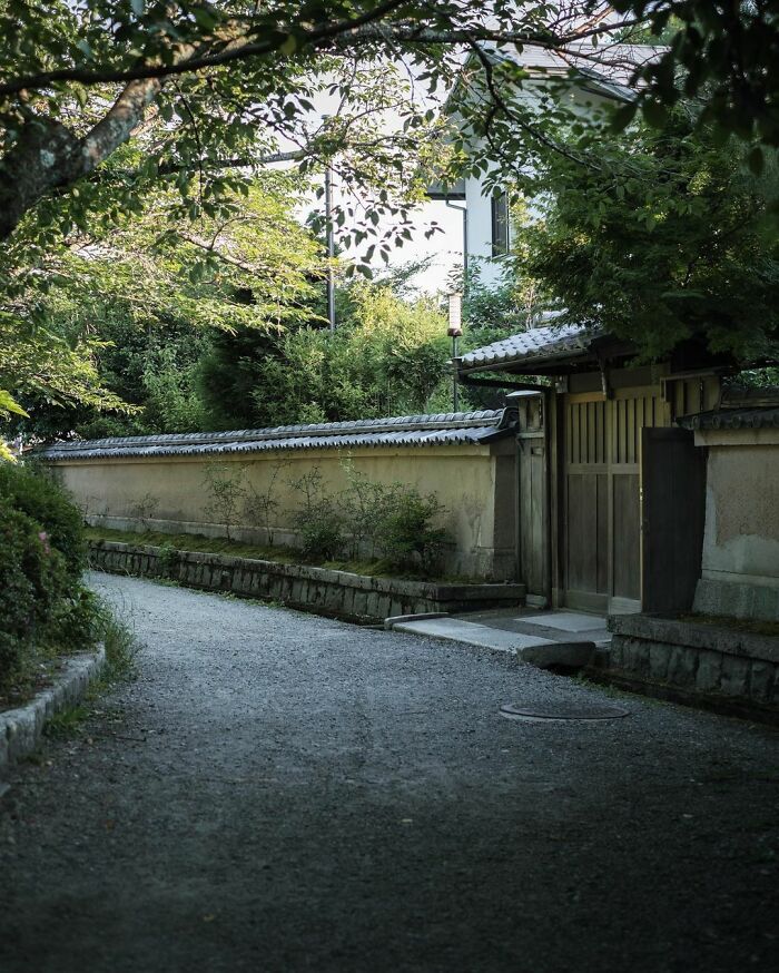 Traditional Japanese old architecture with wooden gate and stone walls surrounded by greenery along a narrow gravel path.