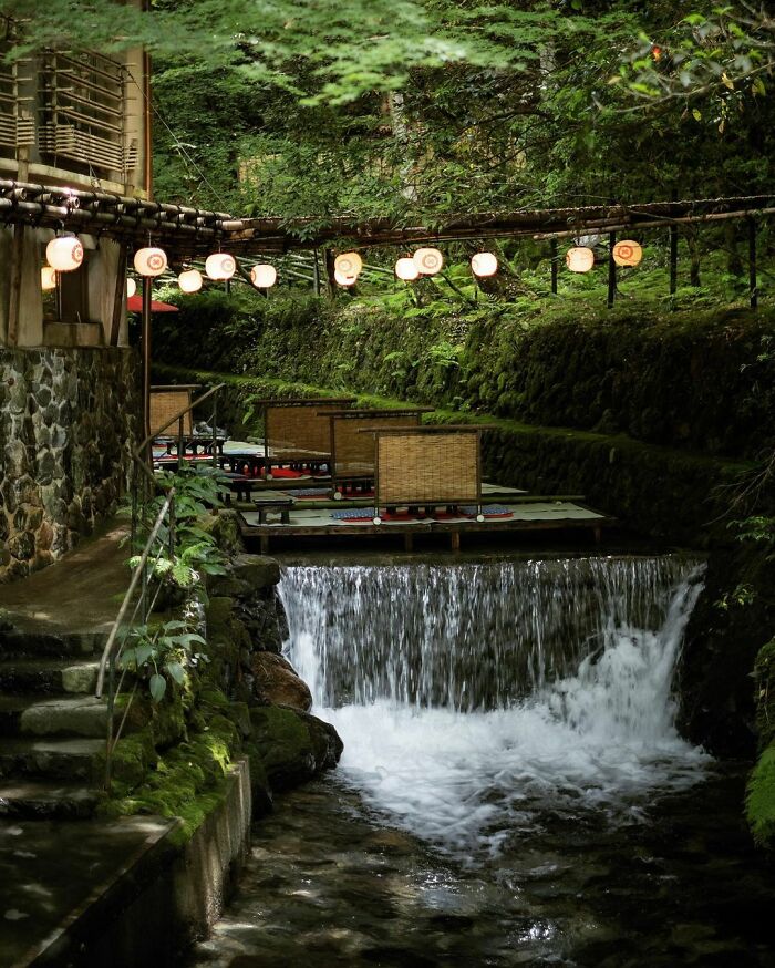 Old architecture in Japan featuring traditional riverside seating with lanterns and lush greenery near a small waterfall.
