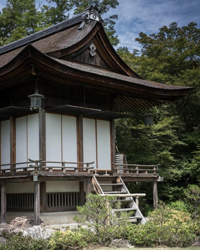 Traditional old architecture in Japan featuring wooden structure, sliding doors, and a thatched roof surrounded by lush greenery.
