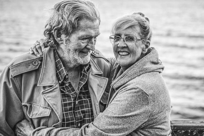 Elderly couple in love smiling and embracing near the water, capturing genuine moments of couples in love photography.
