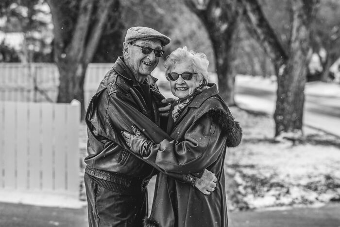 Elderly couple in love smiling and embracing outdoors wearing coats and sunglasses in a black and white photo.