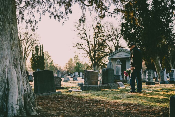 Man wearing hat holding flowers stands in cemetery surrounded by tombstones, illustrating disturbing facts to ruin your day.