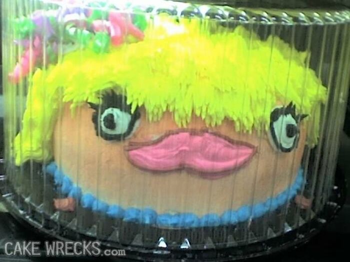 Decorated cake with humorous face; bright yellow hair, bold eyes, pink lips, enclosed in a clear plastic container. Cake fails example.