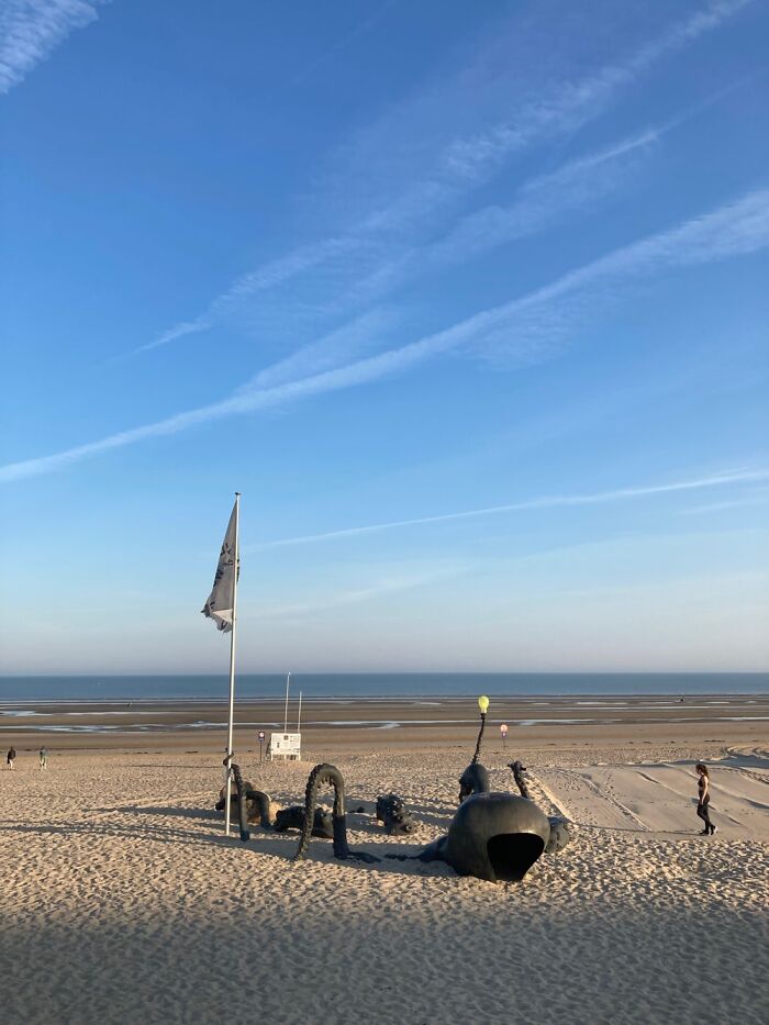 Sandy beach under clear sky with people and a large sculpture during calm weather in September.