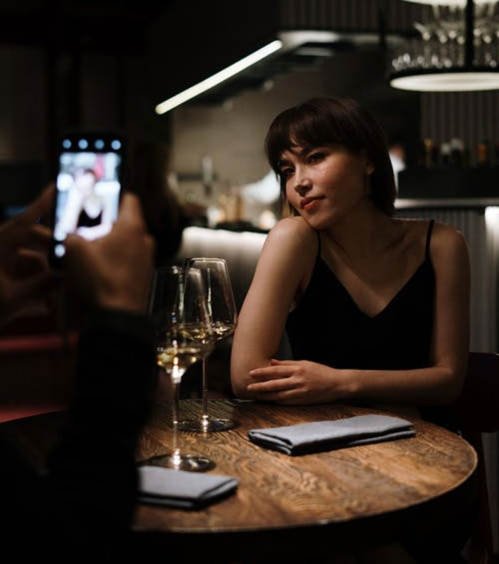 A woman posing at a restaurant table with wine glasses as someone takes her photo, highlighting pretty privilege issues.