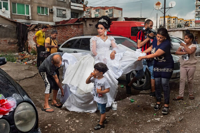 Bride in an elaborate white dress is helped by people in a rough urban setting, showcased in Siena International Photo Awards.