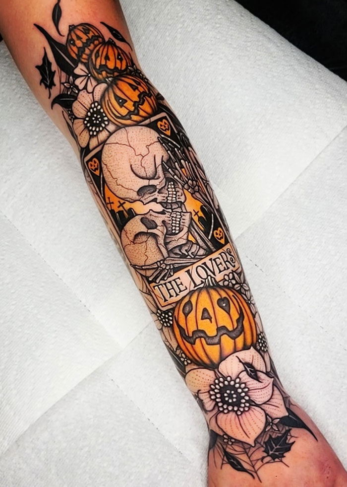 Spooky Halloween tattoo featuring skulls, pumpkins, and flowers on a person's forearm.