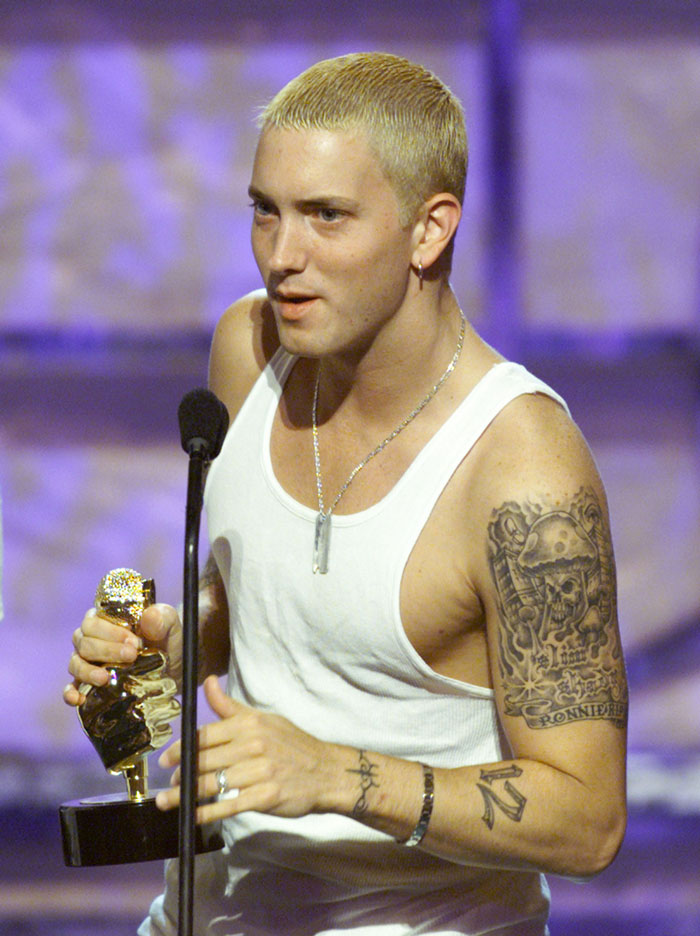 Person holding an award on stage, wearing a white tank top. Nostalgic 2000s fashion and tattoo visible.
