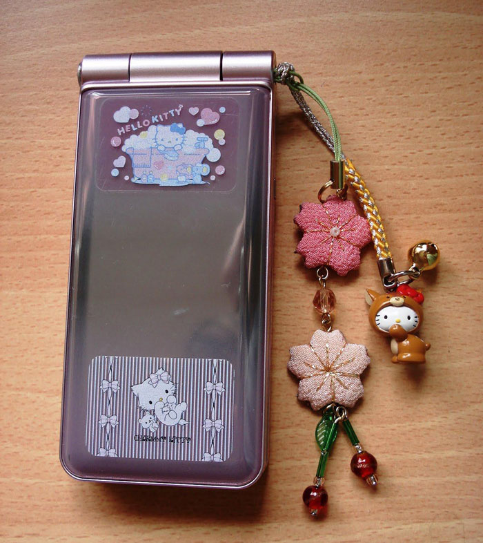 Pink flip phone with Hello Kitty design, decorated with nostalgic 2000s charms featuring flowers and a character.