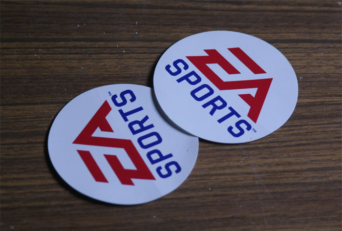 Two round stickers with EA Sports logo on a wooden surface, illustrating sarcastically honest slogans about famous brands.