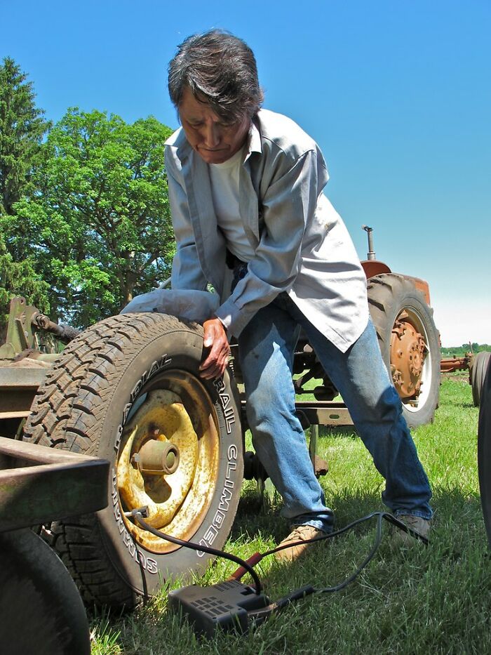 Person using a portable air compressor to inflate a tractor tire outdoors in a sunny, grassy area.