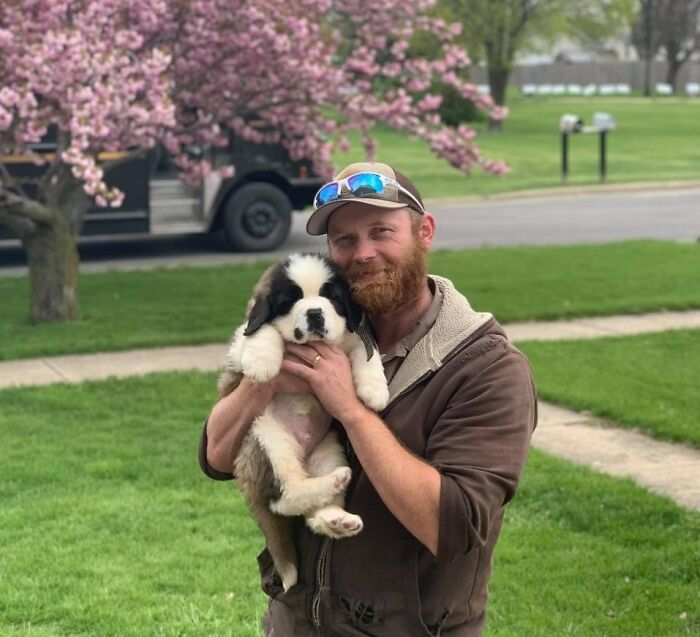 UPS driver holding a puppy outside with a UPS truck and blooming tree in the background during a delivery route.