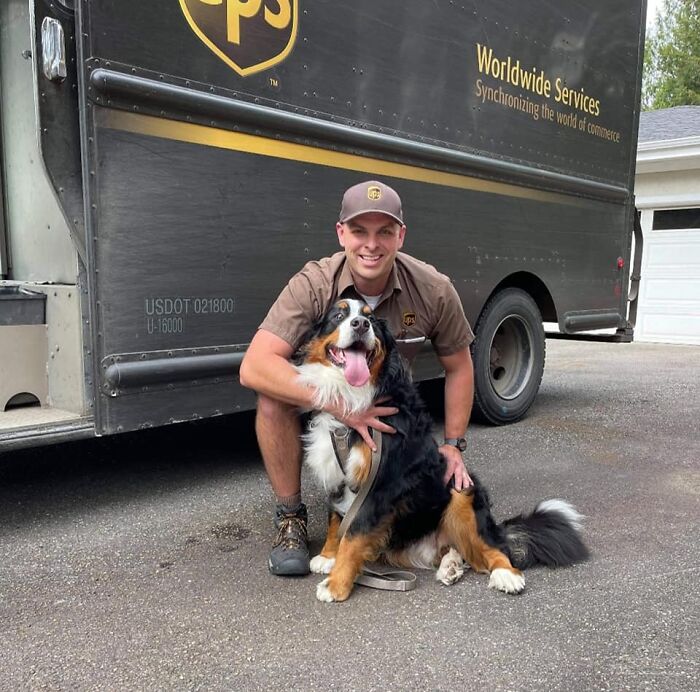 UPS driver in uniform kneeling and smiling while hugging a large happy dog in front of a UPS delivery truck.