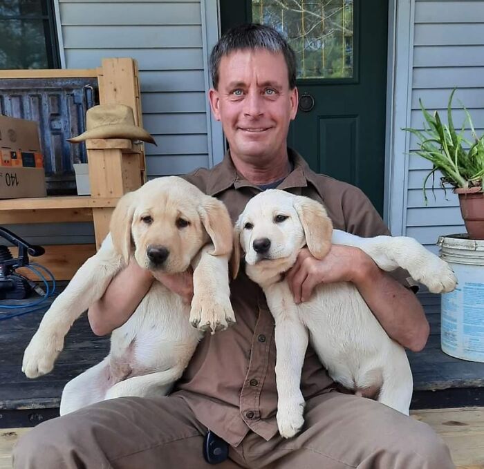 UPS driver sitting on porch holding two Labrador puppies he met on his delivery route smiling warmly