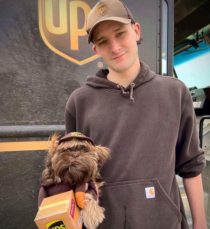 UPS driver holding a dog dressed in a UPS uniform, posing in front of a UPS delivery truck on his route.
