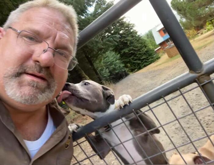 UPS driver taking a selfie while a dog licks his face over a gate on his delivery route in a rural area.
