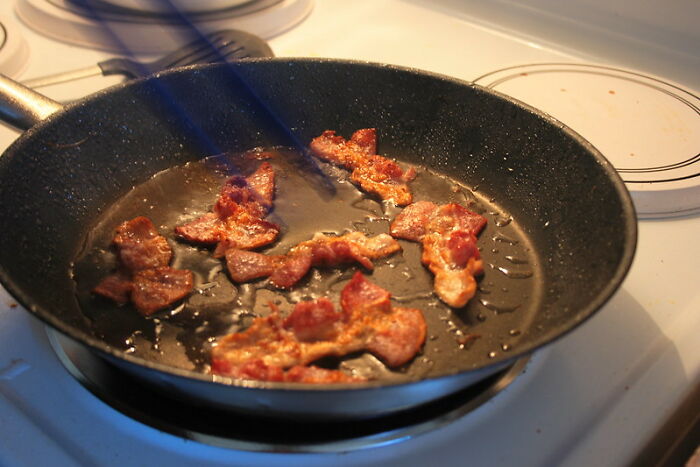 Crispy bacon sizzling in a black frying pan on a white electric stove top, cooking breakfast food product.