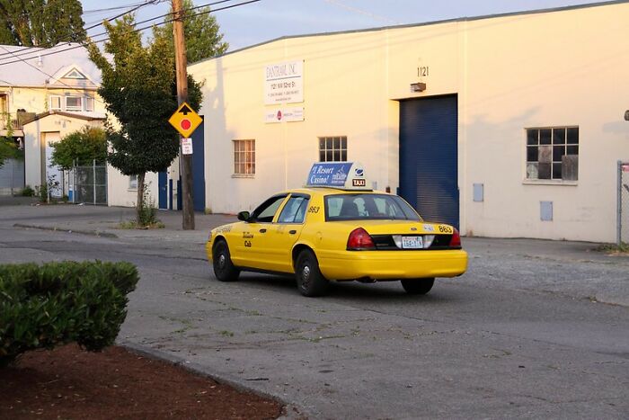 Yellow taxi driving on a quiet urban street near industrial buildings in late afternoon light.