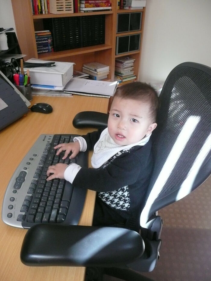 Toddler sitting in office chair using a keyboard at a desk with books and stationery in an online group setting.