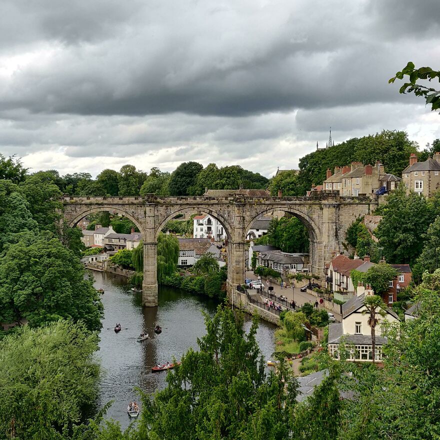 Historic stone viaduct spanning a river, surrounded by lush greenery and charming village scenery.