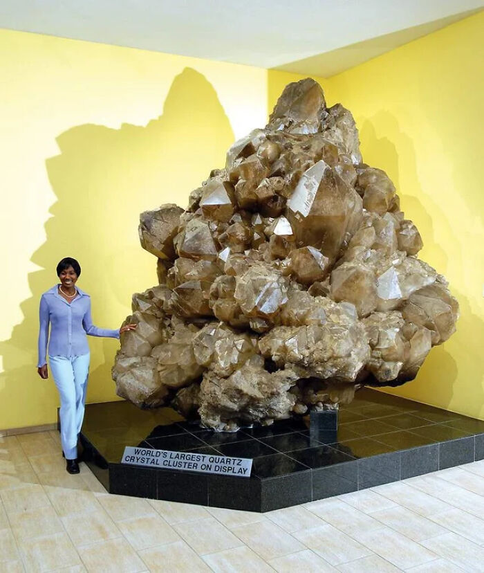 Woman standing next to an absolute unit quartz crystal cluster, the world's largest on display in a museum setting.